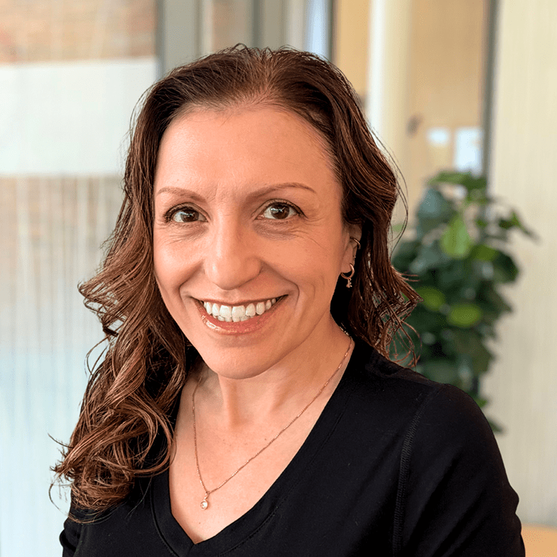 Smiling woman with wavy brown hair wearing a black top and a necklace, standing indoors with a blurred green plant and window in the background. Amy L. Brandt, DNP, APRN, exudes warmth and professionalism in this inviting setting.