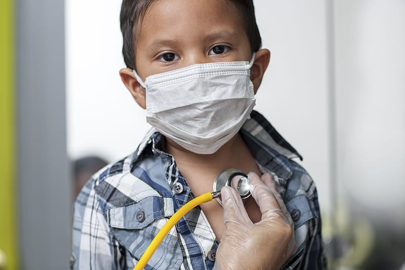 A young boy wearing a plaid shirt and a face mask is being examined with a stethoscope by a gloved healthcare professional, who is carefully providing important health information, possibly in Spanish, to ensure clear communication with the patient.