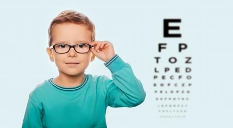 Young child wearing glasses and a teal shirt stands in front of an eye chart, touching their glasses, with a light blue background—perfect for highlighting the latest Fall Eye Sale.