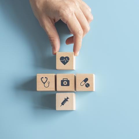 A hand arranges wooden blocks with medical icons—such as a heart, stethoscope, first aid kit, pill, and syringe—on a light blue background, highlighting health as a priority.