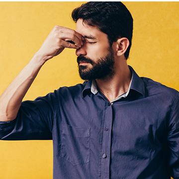 A man with a beard, wearing a dark shirt, stands against a yellow background, pinching the bridge of his nose with his eyes closed, appearing stressed or fatigued—possibly struggling with allergy symptoms.