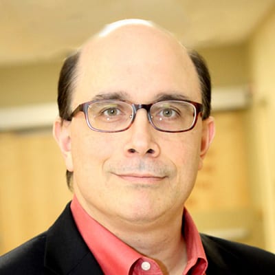 A middle-aged man with glasses, short dark hair, and a receding hairline, wearing a red collared shirt and black blazer, stands indoors with a blurred background.