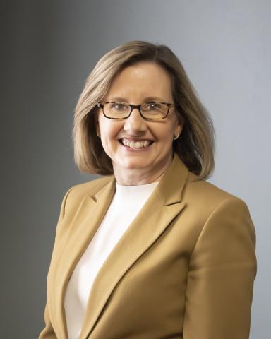 A woman with shoulder-length blonde hair, wearing glasses, a beige blazer, and a white top, smiles at the camera against a plain gray background—an approachable and professional depiction of Kerrie McEvilly, president of SCMC.