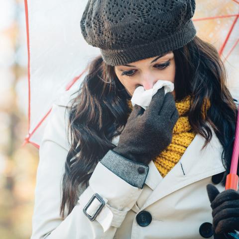 A woman wearing a coat, knit hat, and gloves holds an umbrella and uses a tissue to wipe her nose, suggesting she might be experiencing allergy symptoms.