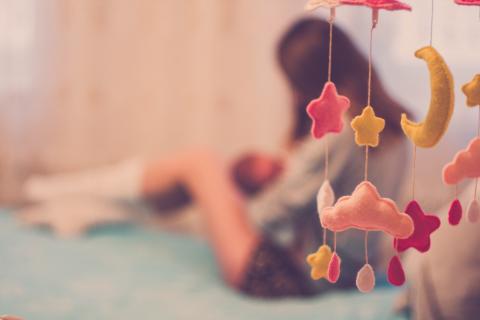 A soft-focus image shows a woman breastfeeding a baby on a bed, with colorful felt stars, clouds, and a moon mobile hanging in the foreground.