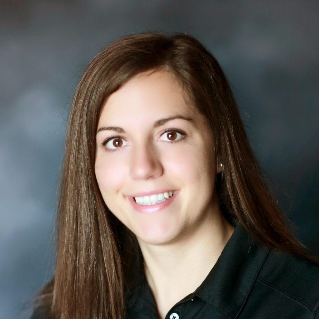Smiling woman with long brown hair, wearing a black collared shirt, posed against a blurred dark background.