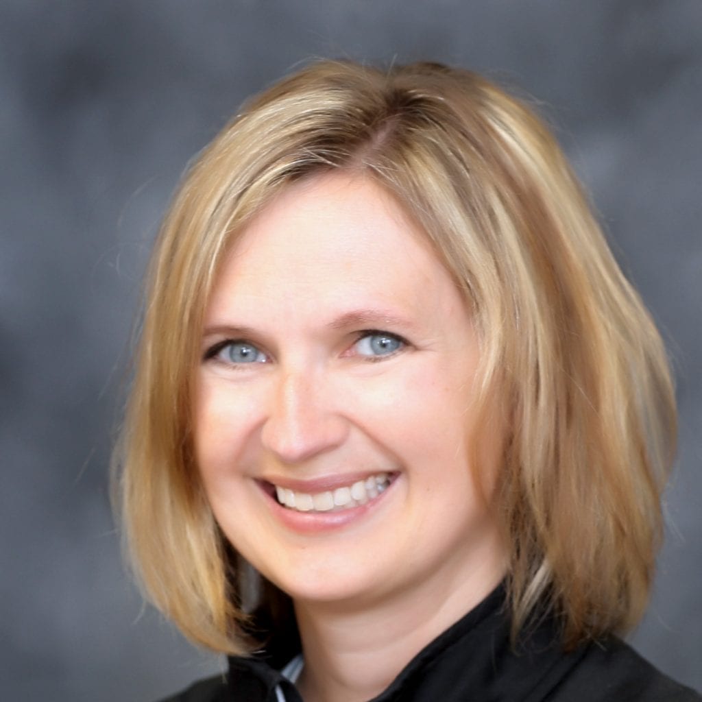 Smiling woman with straight, shoulder-length blonde hair, wearing a black top, posing in front of a blurred gray background.