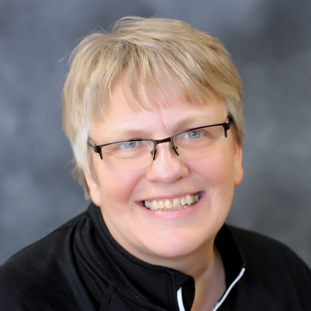 A person with short blonde hair, wearing glasses and a black zip-up top, smiles at the camera against a blurred gray background.