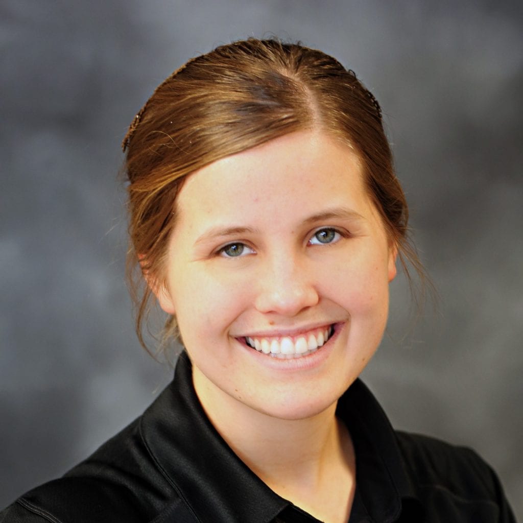 A young woman with light brown hair in a neat bun, wearing a black collared shirt, is smiling at the camera against a blurred gray background.