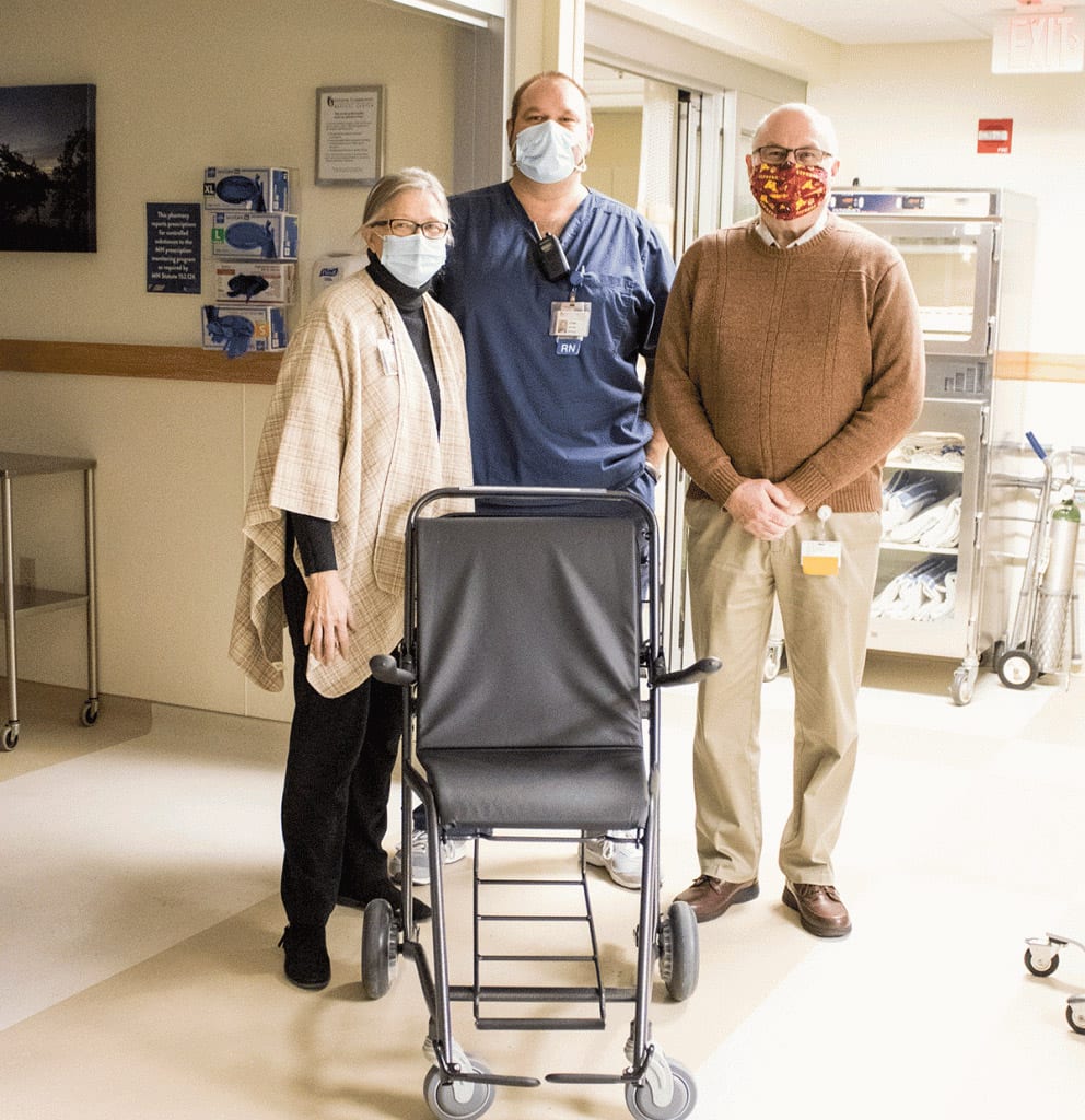 Three adults wearing masks stand in a hospital hallway behind a black wheelchair. Two wear casual clothes and one wears blue medical scrubs. Medical equipment and supplies are visible in the background.