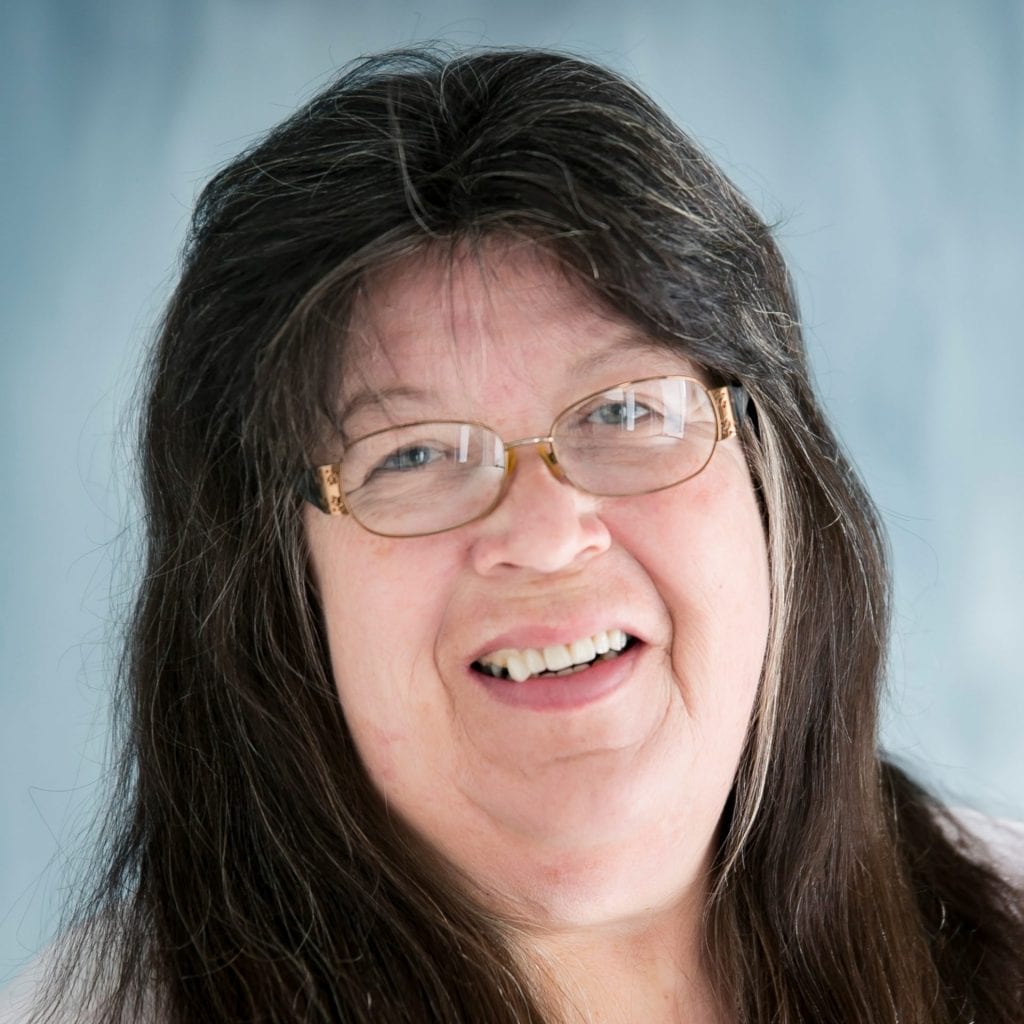 Smiling older woman with long dark hair and glasses, wearing a light-colored top, in front of a soft blue background.