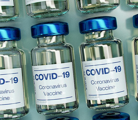 Several glass vials with blue caps are labeled “COVID-19 Coronavirus Vaccine” and are arranged in rows against a light background, highlighting the vital role vaccines play in combating COVID-19.