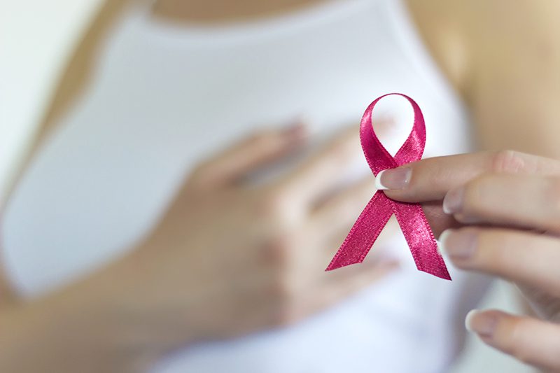 A person in a white tank top holds a pink ribbon, a symbol of breast cancer awareness, with one hand while placing the other hand on their chest.