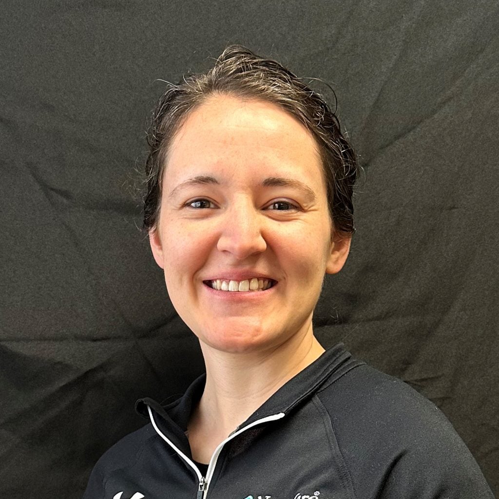 A person with short, dark hair smiles at the camera, wearing a black zip-up jacket, with a plain black background behind them.
