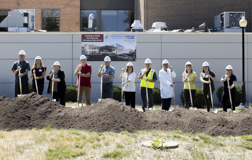 A group of people in hard hats and safety vests stand in a line, holding shovels at a dirt groundbreaking ceremony for an ER expansion in front of a building with a sign about a new emergency department.