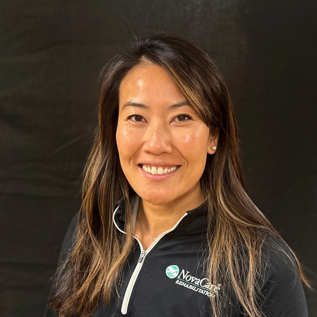 Elizabeth Jenson, DPT, with long brown hair smiling, wearing a black zip-up top with the “NovaCare Rehabilitation” logo, stands in front of a plain dark background.