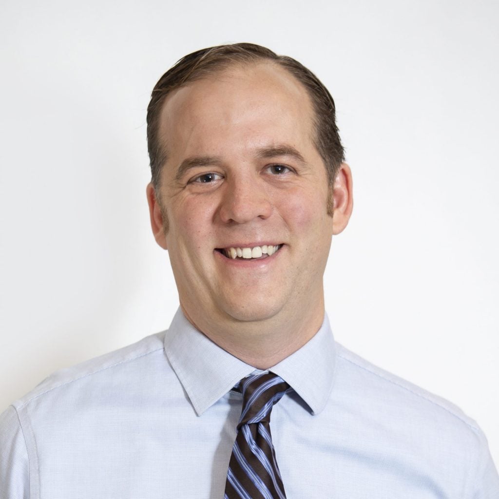 A man in a light blue dress shirt and striped tie smiles at the camera against a plain white background, introducing himself as Darrin J. Spoden, DO.