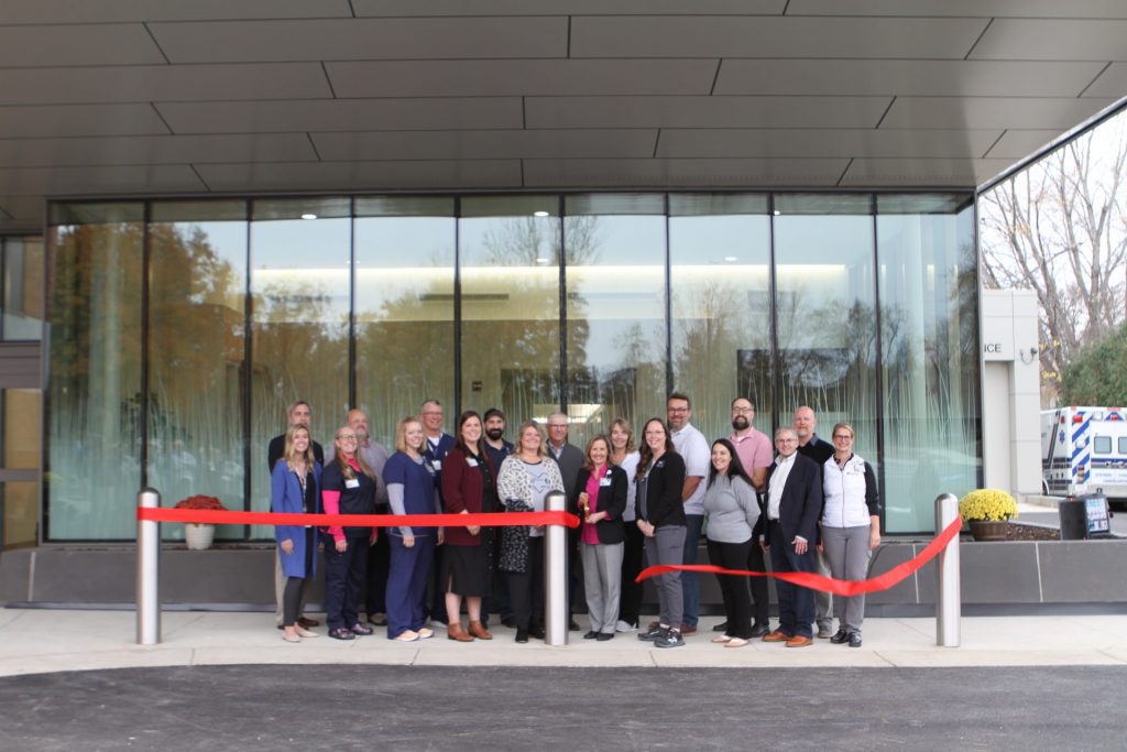 A group of people poses outside a building behind a red ribbon, appearing to celebrate a ribbon-cutting event for an Open House. The background features large glass windows and an ambulance parked to the right.
