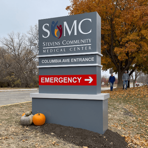 Outdoor sign for Stevens Community Medical Center with directions to the Columbia Ave entrance and Emergency room. Three pumpkins sit at the base; trees with orange leaves and people walking in the background, hinting at the excitement of an upcoming Open House and Ribbon-Cutting event.