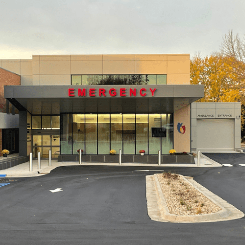 Modern hospital emergency entrance with large red “EMERGENCY” sign above glass doors, ambulance entrance sign to the right, and a blacktop driveway in front. Trees with fall foliage are visible in the background, highlighting SCMC’s ongoing Emergency Dept. Expansion and recent upgrades to improve patient care and accessibility.