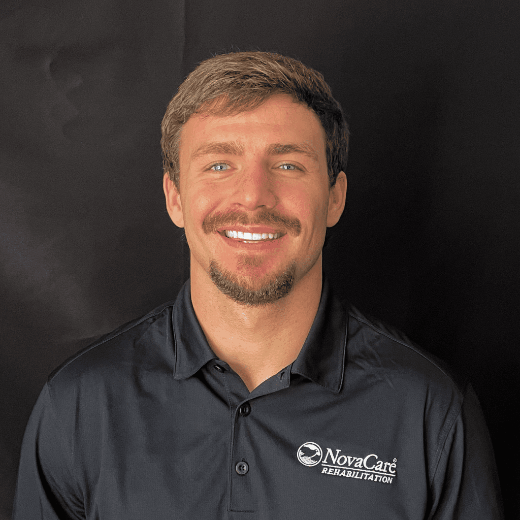 Young man with short light brown hair and goatee, wearing a black NovaCare Rehabilitation polo shirt, smiling in front of a plain black background.