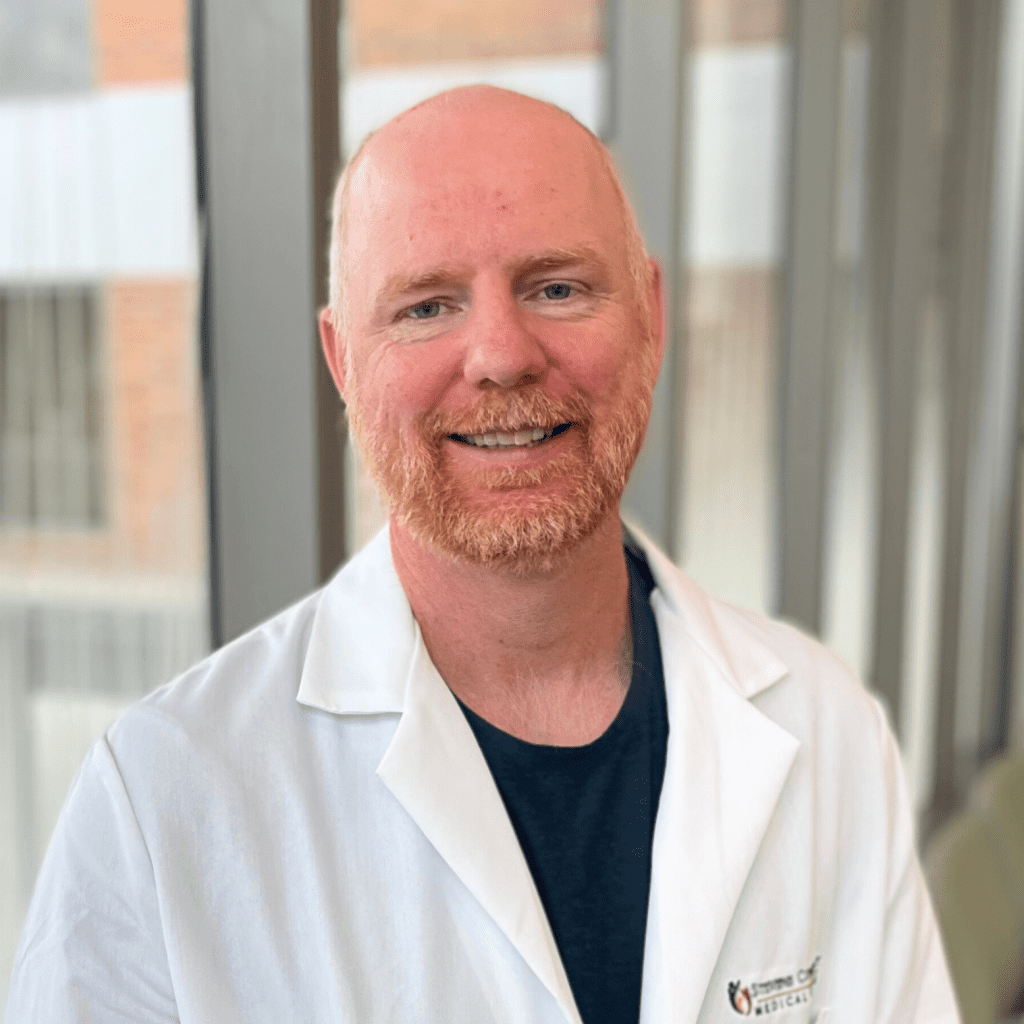 A man with a reddish beard, wearing a white lab coat over a dark shirt, smiles while sitting indoors near large windows with blurred outdoor buildings in the background.