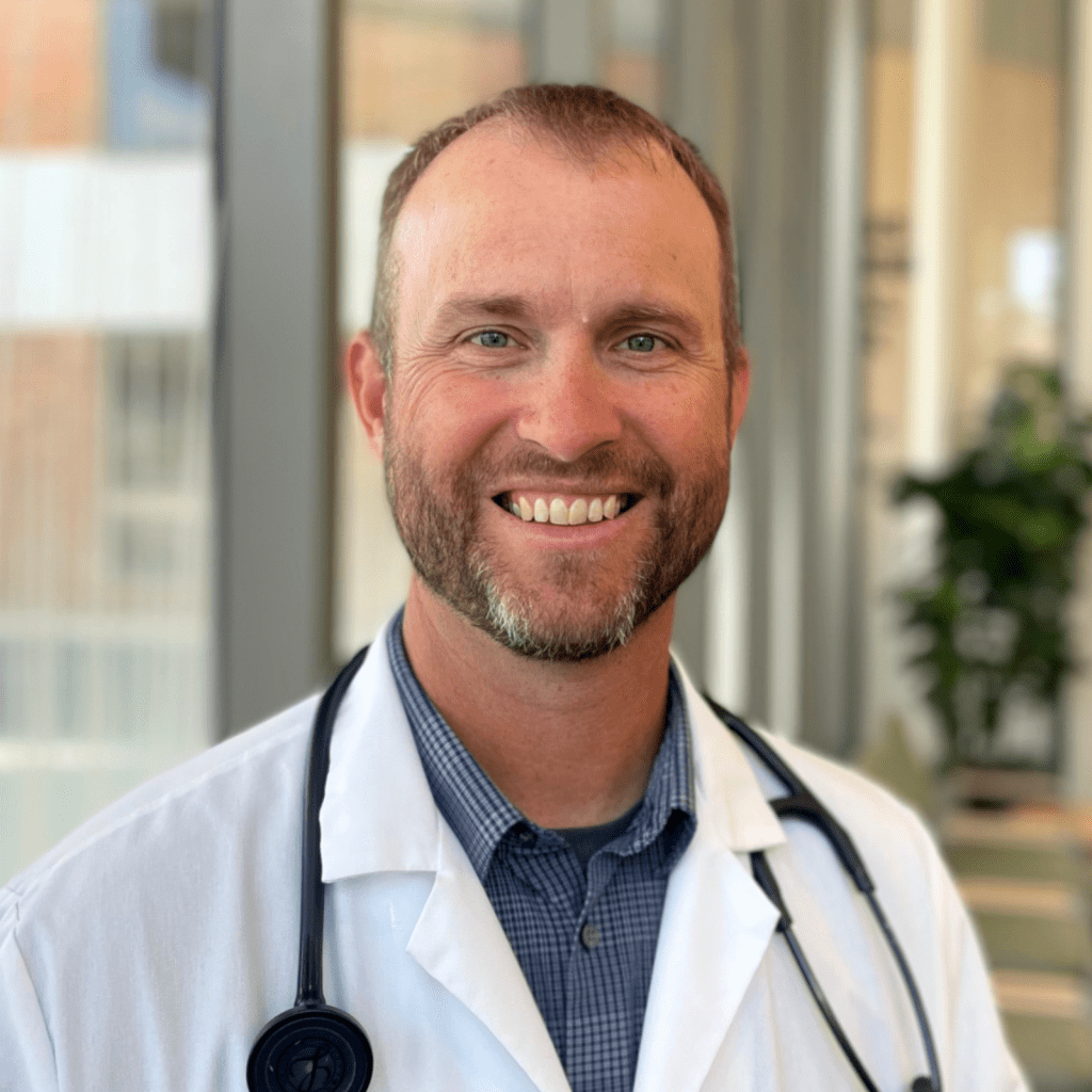 A smiling man with a beard, wearing a white lab coat and a stethoscope, stands indoors with blurred windows and a plant in the background.