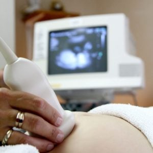A hand holding an ultrasound probe on a patient’s abdomen, with a blurred ultrasound monitor in the background displaying a scan image. The patient is covered with a white towel.