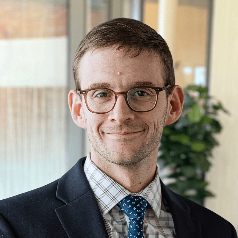 A man wearing glasses, a dark suit, a plaid shirt, and a blue patterned tie smiles in an office setting with a plant and windows in the background. This is Samuel J. Bloomsburg, MD.