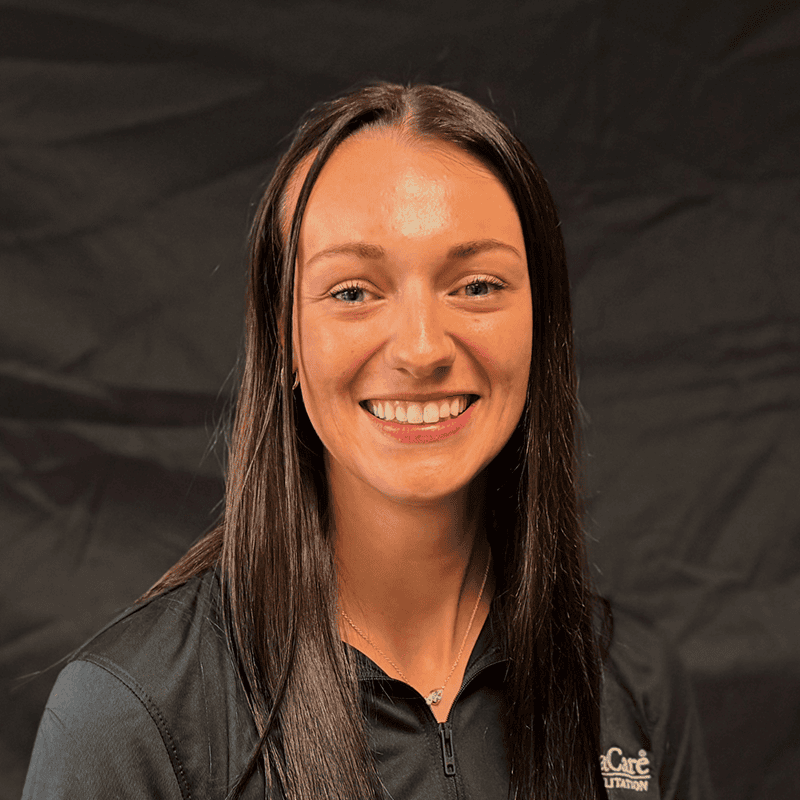 A young woman with long brown hair smiles at the camera, wearing a black collared shirt. The background is plain black.