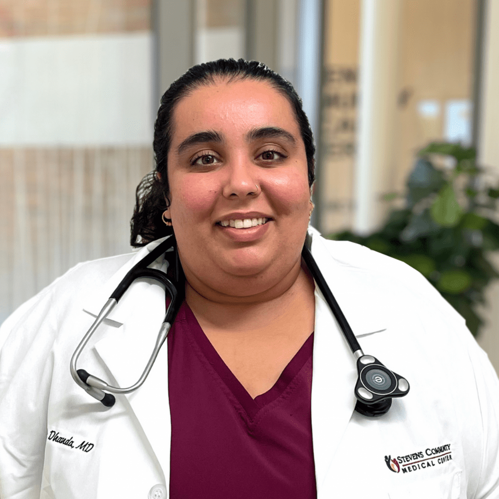A doctor wearing a white coat and stethoscope smiles at the camera, standing indoors in a medical facility with plants and glass in the background.