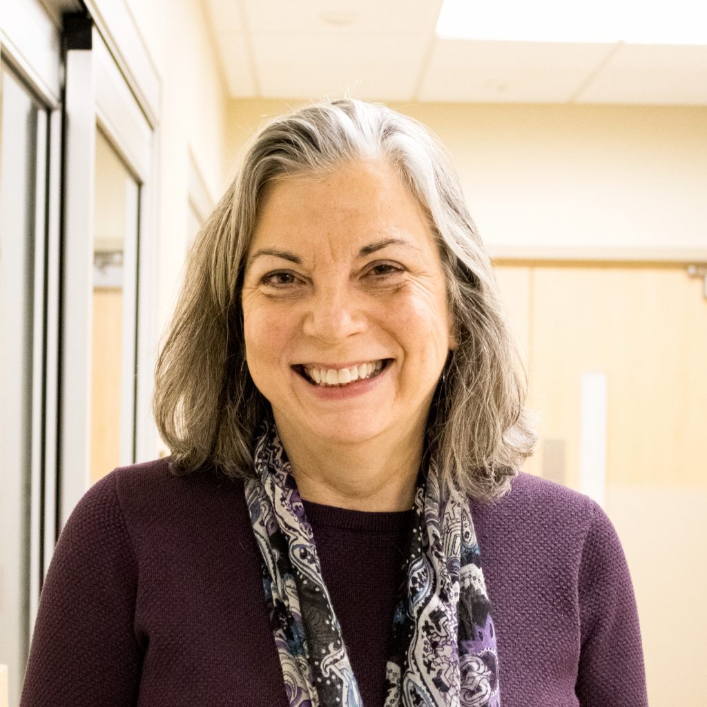 A woman with gray hair, wearing a purple sweater and patterned scarf, smiles while standing indoors in a well-lit hallway.