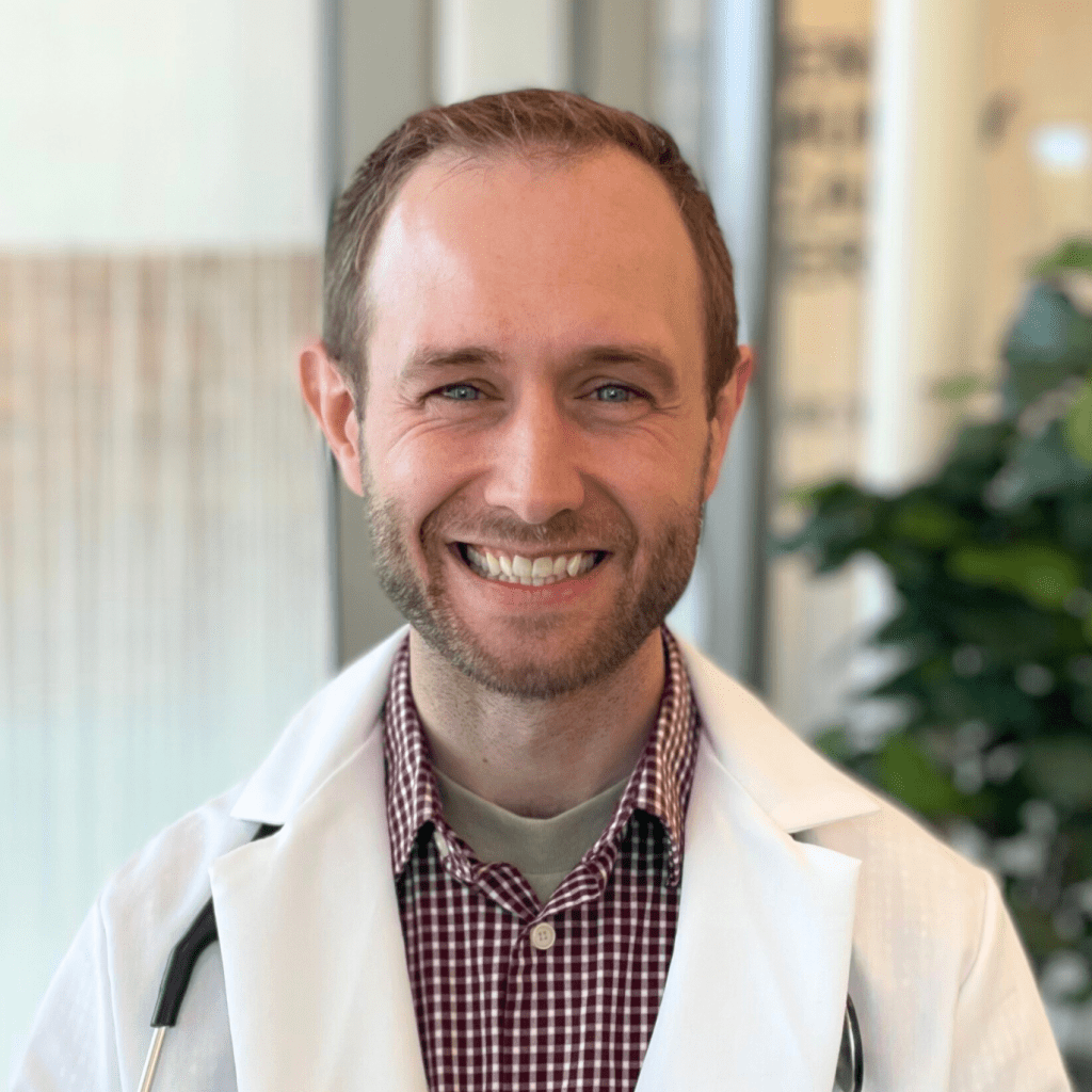 A man in a white lab coat and checkered shirt smiles at the camera, standing indoors with a plant and window in the background.
