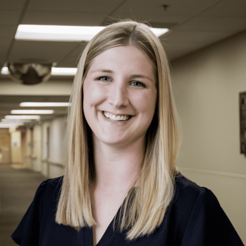 A woman with straight blonde hair, wearing a dark top, smiles in a well-lit hallway with soft beige walls and ceiling lights—this is Tierza Thormodson, a dedicated nurse practitioner with her DNP.